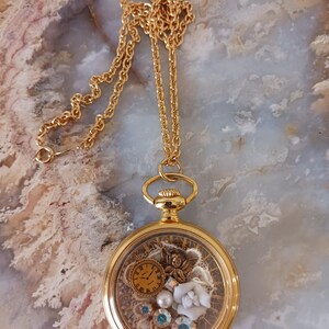 Puede incluir: Collar con colgante de reloj de bolsillo dorado. La esfera del reloj es transparente, revelando un reloj en miniatura, un &aacute;ngel, perlas y cristales. La cadena es una delicada cadena dorada.