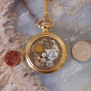 Puede incluir: Colgante de reloj de bolsillo dorado con un interior decorativo que presenta un reloj, un &aacute;ngel, una rosa blanca y cristales iridiscentes. El colgante cuelga de una cadena dorada. Una moneda de un centavo y un cuarto de d&oacute;lar est&aacute;n al lado del colgante.