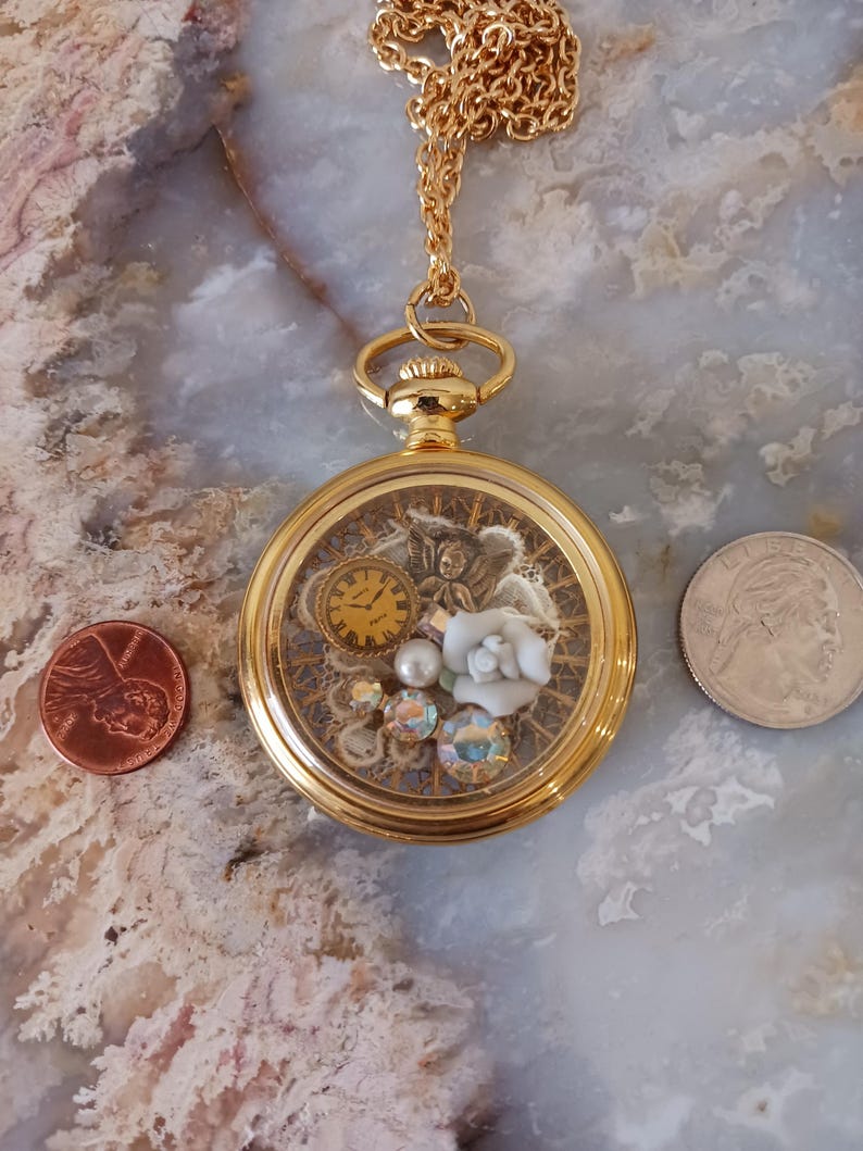 Puede incluir: Colgante de reloj de bolsillo dorado con un interior decorativo que presenta un reloj, un &aacute;ngel, una rosa blanca y cristales iridiscentes. El colgante cuelga de una cadena dorada. Una moneda de un centavo y un cuarto de d&oacute;lar est&aacute;n al lado del colgante.