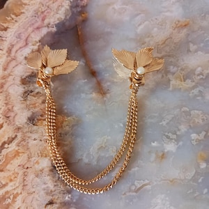 May include: Gold-toned collar clip with a floral design. Each clip features a detailed flower with a small pearl in the center, connected by two delicate gold chains. The clips are displayed on a light-colored, marbled surface.