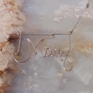 May include: A delicate silver wire necklace spelling out the name "Mary" in cursive script. The wire is shaped into the letters and suspended on a thin chain. The necklace is displayed against a marbled background.
