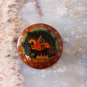 May include: Round, hand-painted wooden trinket box with a vibrant scene of a figure on horseback. The box features a dark red border with gold floral accents and a black interior with a castle and trees. The rider is dressed in red and gold.