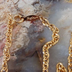 Puede incluir: Primer plano de un collar dorado con un dise&ntilde;o de eslabones de cadena. El cierre est&aacute; marcado con la marca "Napier". El collar se muestra sobre un fondo moteado de tonos neutros.