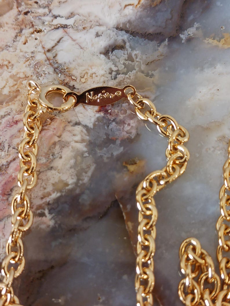 Puede incluir: Primer plano de un collar dorado con un dise&ntilde;o de eslabones de cadena. El cierre est&aacute; marcado con la marca "Napier". El collar se muestra sobre un fondo moteado de tonos neutros.