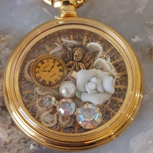 Puede incluir: Un colgante de reloj de bolsillo dorado con una esfera transparente, que muestra un reloj en miniatura, una figura de &aacute;ngel, una rosa blanca, perlas y cristales iridiscentes. El reloj tiene una cadena y un cierre dorados decorativos.