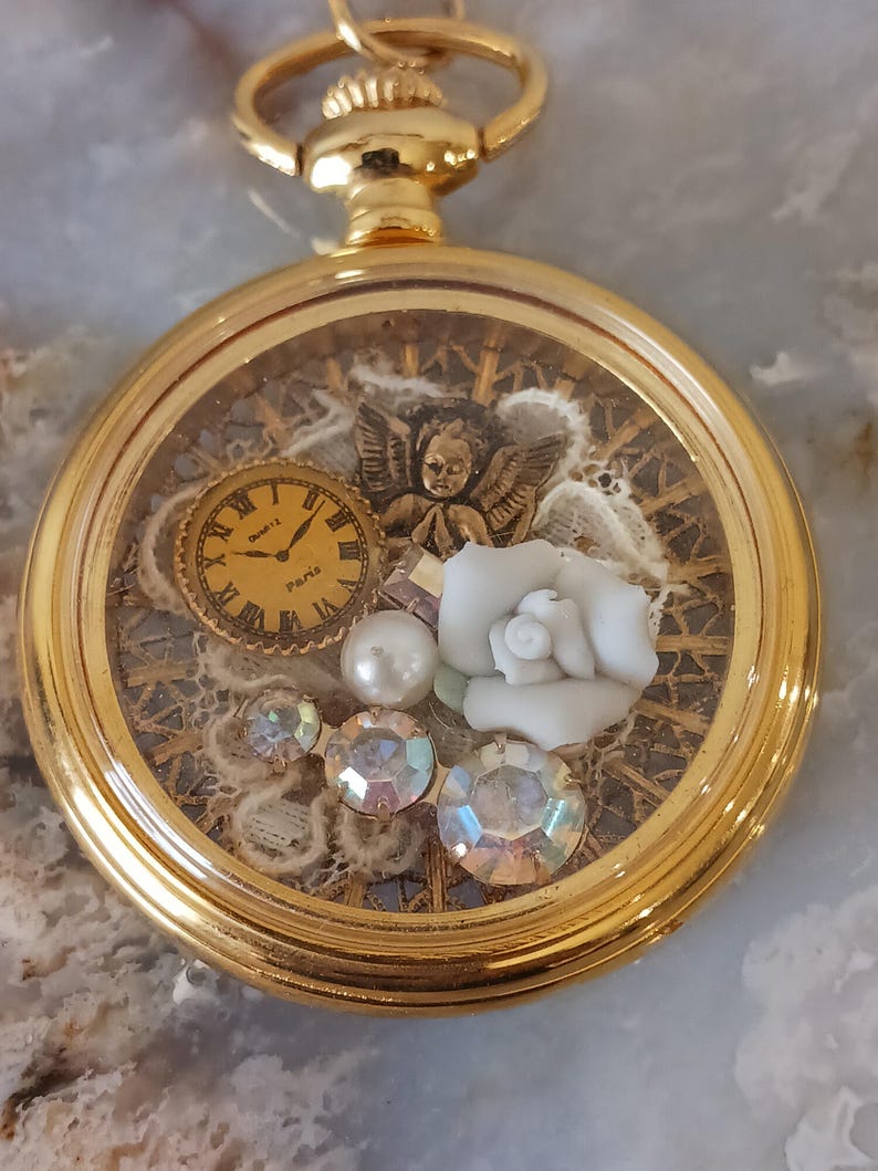 Puede incluir: Un colgante de reloj de bolsillo dorado con una esfera transparente, que muestra un reloj en miniatura, una figura de &aacute;ngel, una rosa blanca, perlas y cristales iridiscentes. El reloj tiene una cadena y un cierre dorados decorativos.