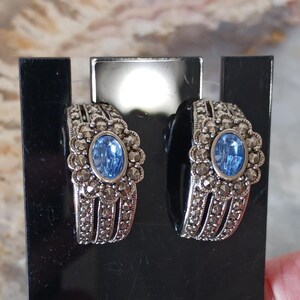 May include: A pair of silver-toned earrings with a marcasite design. Each earring features a central oval-shaped, light blue gemstone surrounded by a floral-like setting. The earrings are displayed on a black stand.