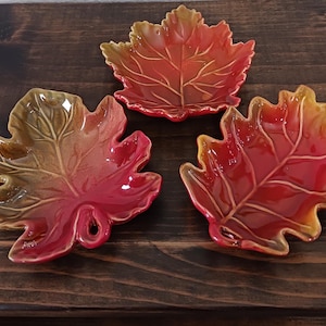 May include: Three ceramic leaf-shaped dishes in autumnal colors. The dishes are glazed in shades of red, orange, and brown, with detailed veining. The dishes are arranged on a dark wooden surface, perfect for fall decor.