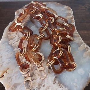 May include: A brown acrylic chain with gold-colored metal accents. The chain is composed of large, rectangular links and features two gold-colored clasps. The chain is displayed on a light-colored, patterned surface.