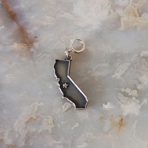May include: Silver charm in the shape of the state of California with a small star in the center. The charm has a silver clasp.