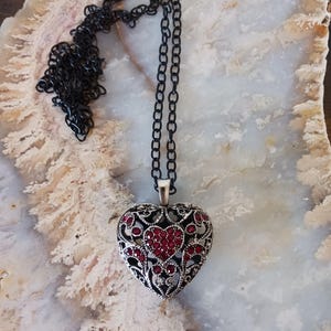 May include: A silver-coloured heart pendant necklace with a black chain. The heart pendant has an openwork design with red accents. The necklace is displayed on a light-coloured, patterned surface.