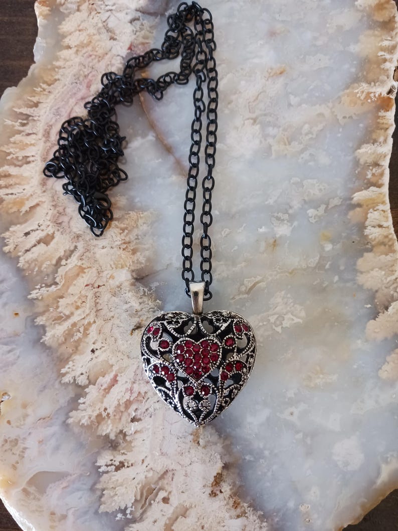 May include: A silver-coloured heart pendant necklace with a black chain. The heart pendant has an openwork design with red accents. The necklace is displayed on a light-coloured, patterned surface.