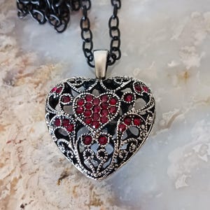 May include: A silver-toned heart pendant necklace with a black chain. The heart pendant features a detailed filigree design and is embellished with red gemstones. The necklace is a piece of jewellery.
