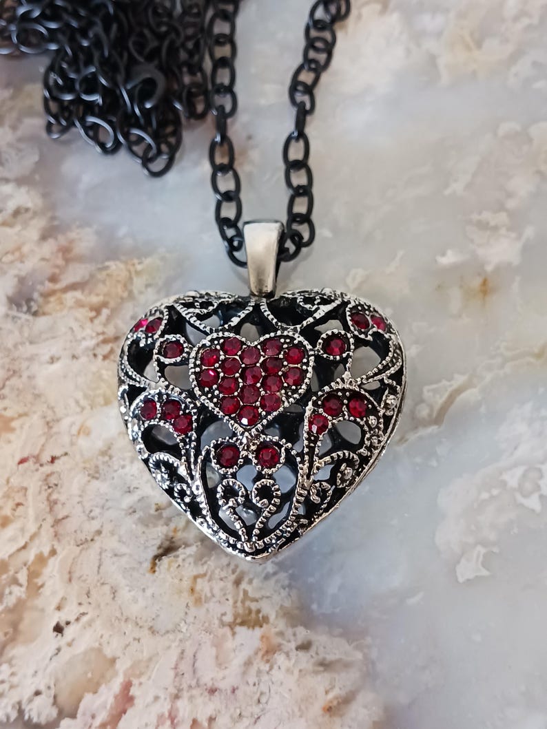 May include: A silver-toned heart pendant necklace with a black chain. The heart pendant features a detailed filigree design and is embellished with red gemstones. The necklace is a piece of jewellery.