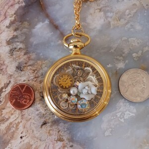 Puede incluir: Collar con medall&oacute;n dorado con detalles intrincados en el interior. El medall&oacute;n contiene un peque&ntilde;o reloj, una rosa blanca, cristales iridiscentes y una perla. La cadena tambi&eacute;n es dorada. Una moneda y un cuarto de d&oacute;lar est&aacute;n al lado.