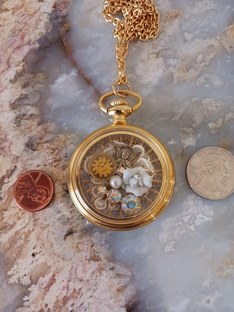 Puede incluir: Collar con medall&oacute;n dorado con detalles intrincados en el interior. El medall&oacute;n contiene un peque&ntilde;o reloj, una rosa blanca, cristales iridiscentes y una perla. La cadena tambi&eacute;n es dorada. Una moneda y un cuarto de d&oacute;lar est&aacute;n al lado.
