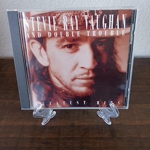 May include: A CD case featuring Stevie Ray Vaughan and Double Trouble's "Greatest Hits" album. The cover art displays a portrait of Stevie Ray Vaughan in a hat. The CD case is clear, and the album title is prominently displayed.