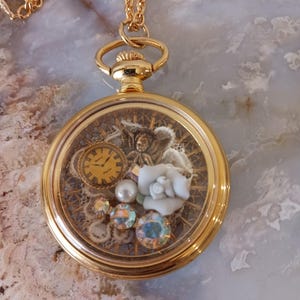 Puede incluir: Collar con colgante de reloj de bolsillo dorado. La esfera del reloj presenta un peque&ntilde;o reloj, una rosa blanca y cristales iridiscentes. El reloj cuelga de una delicada cadena dorada. La caja del reloj tiene aproximadamente 3,8 cm de di&aacute;metro.