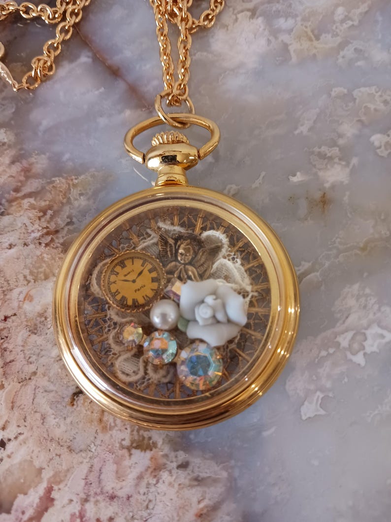 Puede incluir: Collar con colgante de reloj de bolsillo dorado. La esfera del reloj presenta un peque&ntilde;o reloj, una rosa blanca y cristales iridiscentes. El reloj cuelga de una delicada cadena dorada. La caja del reloj tiene aproximadamente 3,8 cm de di&aacute;metro.