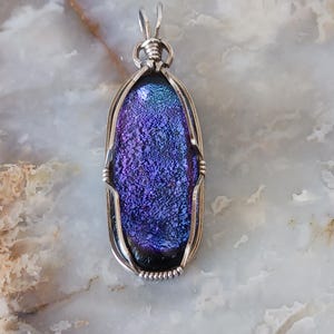 May include: A dichroic glass pendant with a silver wire wrap. The glass has a textured, iridescent appearance with shades of purple, blue, and black. The pendant is oval-shaped and hangs from a silver bail. The pendant is approximately 2 inches long.