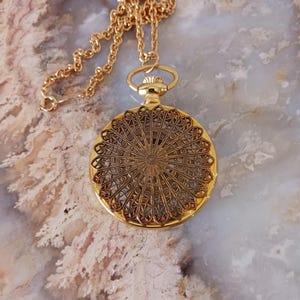 Puede incluir: Un collar con colgante dorado con un dise&ntilde;o de filigrana circular detallado. El colgante cuelga de una delicada cadena. El intrincado patr&oacute;n del colgante es visible, mostrando una est&eacute;tica vintage. El collar se exhibe sobre una superficie de color claro.