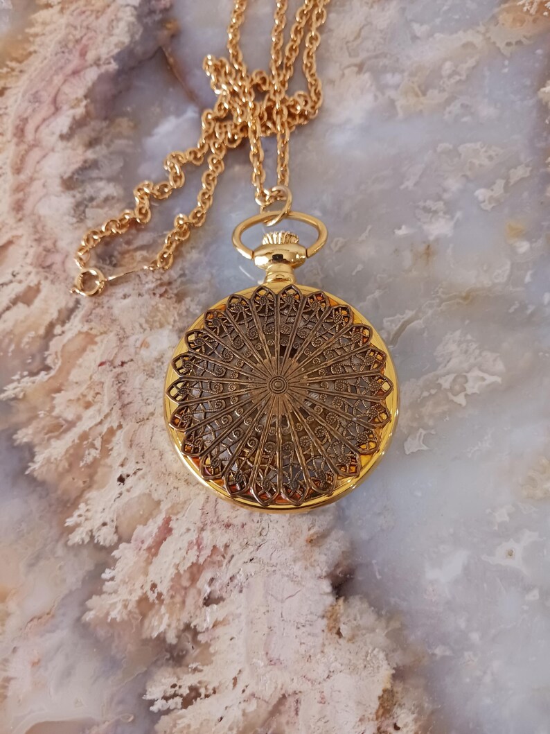 Puede incluir: Un collar con colgante dorado con un dise&ntilde;o de filigrana circular detallado. El colgante cuelga de una delicada cadena. El intrincado patr&oacute;n del colgante es visible, mostrando una est&eacute;tica vintage. El collar se exhibe sobre una superficie de color claro.