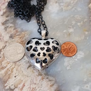 May include: A silver heart-shaped pendant with a black chain. The pendant has heart-shaped cutouts and is displayed with a penny and a coin. The necklace is a piece of jewellery.