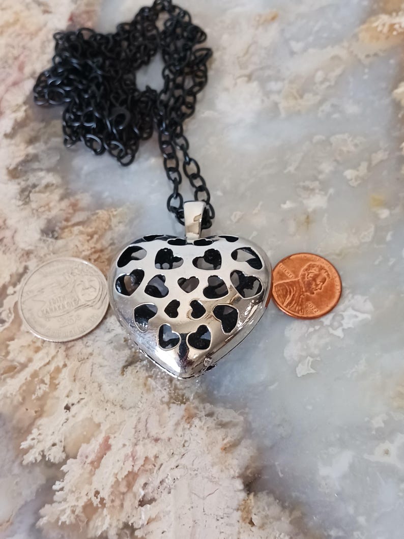 May include: A silver heart-shaped pendant with a black chain. The pendant has heart-shaped cutouts and is displayed with a penny and a coin. The necklace is a piece of jewellery.