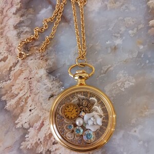 Puede incluir: Collar dorado con un colgante redondo transparente. En el interior, un peque&ntilde;o reloj, una flor blanca y gemas azules y transparentes. La cadena es fina. El di&aacute;metro del colgante es de aproximadamente 3 cm.