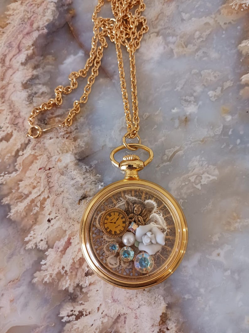 Puede incluir: Collar dorado con un colgante redondo transparente. En el interior, un peque&ntilde;o reloj, una flor blanca y gemas azules y transparentes. La cadena es fina. El di&aacute;metro del colgante es de aproximadamente 3 cm.