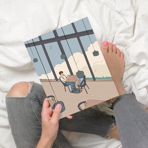 May include: A person is holding a drawing of a couple sitting at a table in a cafe. The cafe has large windows and a view of the sky. The drawing is in a cartoon style.