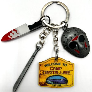 Peut inclure: Un porte-clés argenté avec trois breloques : un couteau ensanglanté, une machette et un panneau Camp Crystal Lake. Le panneau indique "Welcome to Camp Crystal Lake" avec une scène de lac et l'année "EST. 1935". Le masque est gris avec des accents rouges.