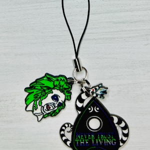 May include: A black and green phone charm with a black and white striped snake design. The charm features a black and white crescent moon and a white circle with the text "NEVER TRUST THE LIVING". The charm is attached to a black string.