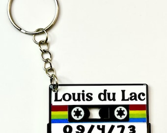 May include: A keychain featuring a flat, rectangular cassette tape charm. The charm is black, white, and features a rainbow stripe. It displays the text 'Louis du Lac' in black lettering and the date '09/4/73' in black font on a white background.