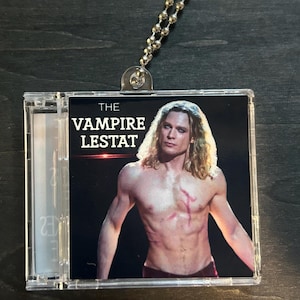 May include: A clear plastic CD case with a silver ball chain. The case features a graphic of a shirtless man with long blonde hair and the text "THE VAMPIRE LESTAT" in white letters.