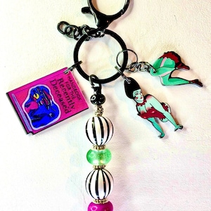 May include: A black keychain with a variety of charms. Charms include a pink book with the text "Handbook for the Recently Deceased", and two cartoon figures. The keychain also has a beaded accent with black and white striped beads, green beads, and pink beads.
