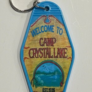 Peut inclure: Un porte-clés bleu de style motel avec les mots "WELCOME TO CAMP CRYSTAL LAKE" dans une police vintage. Le porte-clés présente une scène de lac et le texte "EST. 1935". Le porte-clés est attaché à un anneau en métal argenté.