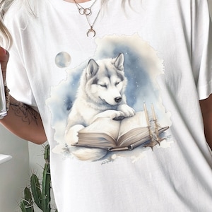 May include: White t-shirt with a watercolor illustration of a wolf reading a book. The wolf is white with gray markings and is sitting on a book with its paws on the pages. The background is a light blue watercolor wash with white stars.