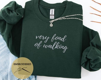 Puede incluir: Sudadera verde bosque con el texto "very fond of walking" bordado en hilo plateado. La sudadera está colocada sobre una superficie blanca con un par de pantalones vaqueros azules y un collar plateado.