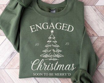 May include: Military green sweatshirt with the text "ENGAGED Christmas" and a Christmas tree graphic. The text "SOON TO BE MERRY'D" is also printed on the sweatshirt. The sweatshirt has a crew neck and long sleeves.