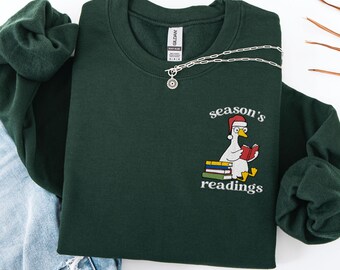 May include: Forest green sweatshirt with the text "season's readings" and an illustration of a goose wearing a Santa hat and reading a book. A silver necklace is draped across the sweatshirt. The sweatshirt is folded on top of a pair of blue jeans.