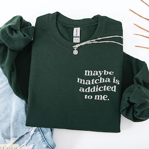 May include: Forest green sweatshirt with the text "maybe matcha is addicted to me." A silver necklace with a pendant is draped across the front. The sweatshirt is folded next to a pair of blue jeans.