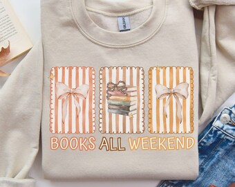 May include: A sand-colored sweatshirt with a crew neck featuring three framed illustrations. The illustrations include bows and a stack of books with the text "BOOKS ALL WEEKEND" in a brown font. The sweatshirt is paired with blue jeans.