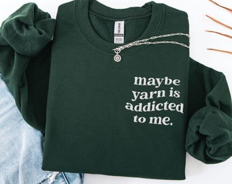 May include: Forest green sweatshirt with the text "maybe yarn is addicted to me." A silver necklace with a pendant is draped across the sweatshirt. The sweatshirt is folded next to a pair of blue jeans.