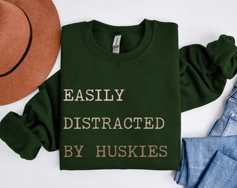 May include: A green sweatshirt with the text "EASILY DISTRACTED BY HUSKIES" printed in a light brown color. The sweatshirt is folded and laid on a white surface with a brown hat and blue jeans.