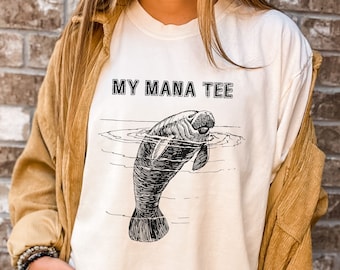 Peut inclure: Un t-shirt blanc à manches longues avec le texte "MY MANA TEE" imprimé en noir. Une illustration en noir et blanc d'un lamantin est imprimée sous le texte.