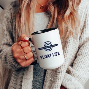 May include: White ceramic mug with a black rim and handle. The mug has a blue graphic of a circle with an arrow and a line through it. The text "FLOAT LIFE" is printed in blue below the graphic.
