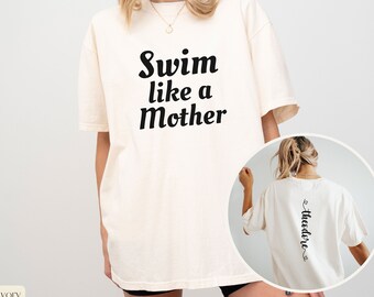 May include: A white oversized t-shirt with the text "Swim like a Mother" printed on the front and the word "Theodore" printed vertically on the back. The shirt is shown on a model.