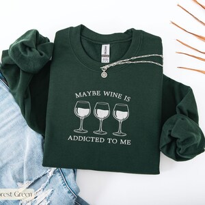May include: Forest green sweatshirt with the text "MAYBE WINE IS ADDICTED TO ME" and three wine glasses. A silver necklace with a pendant is draped across the sweatshirt. The sweatshirt is folded on top of a pair of blue jeans.