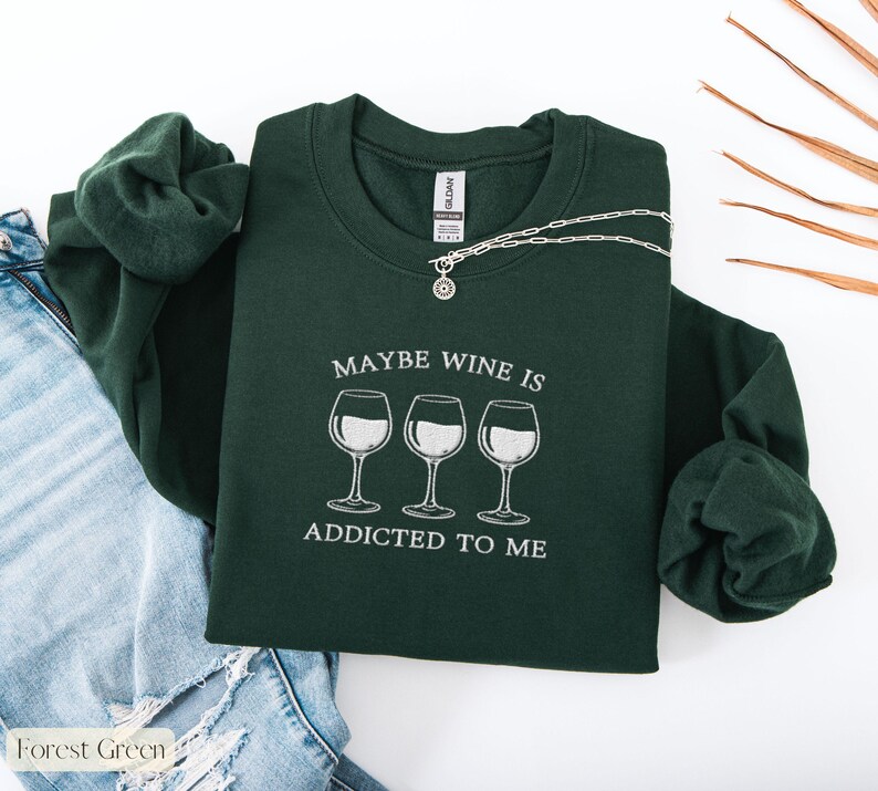 May include: Forest green sweatshirt with the text "MAYBE WINE IS ADDICTED TO ME" and three wine glasses. A silver necklace with a pendant is draped across the sweatshirt. The sweatshirt is folded on top of a pair of blue jeans.